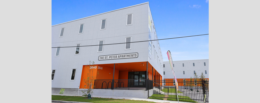 Photograph of the entrance to a three-story apartment building, with a wing of the building in the background.