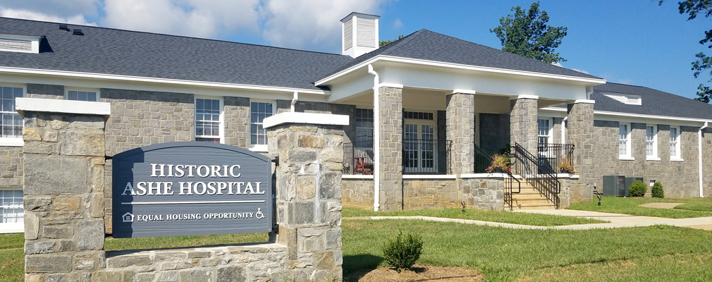 Photograph of the front façade of a stone building with a portico marking the entrance and, in the foreground, a freestanding sign reading “Historic Ashe Hospital.”