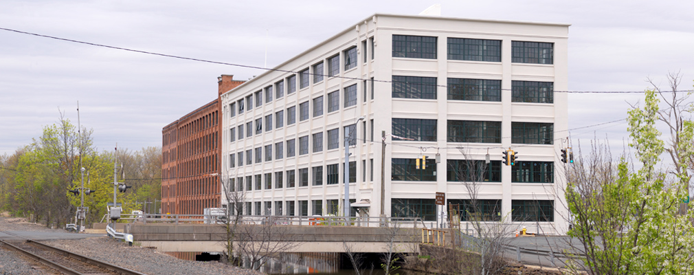 Photograph of two façades of a five-story mill building next to railroad tracks and a canal.
