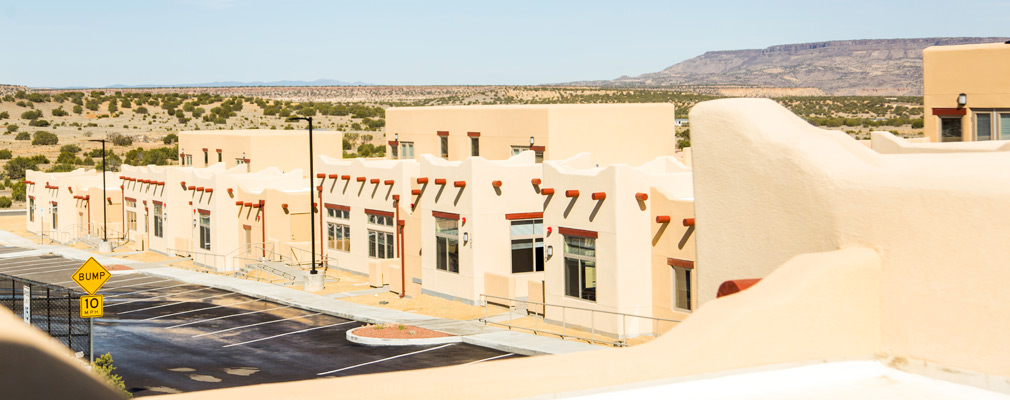 Photograph taken from one story above ground of several one- and two-story attached residences designed in the pueblo style, with a mesa in the background.