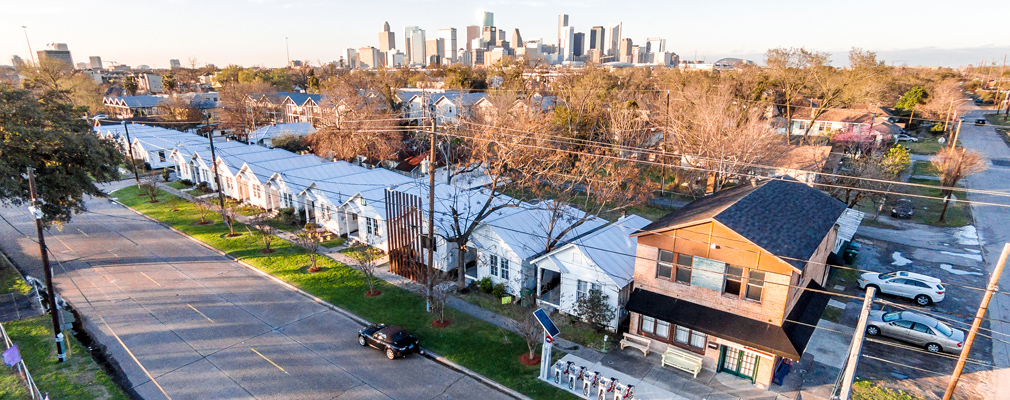 Low-angle aerial photograph of a row of single-story shotgun houses, with a two-story structure to the right of the houses and the skyline of downtown Houston in the background.