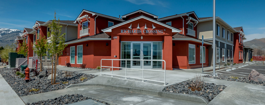 Photograph of the front and side façades of a large two-story building, with several gables accenting the roofline and, in the background, snow-capped mountains. 