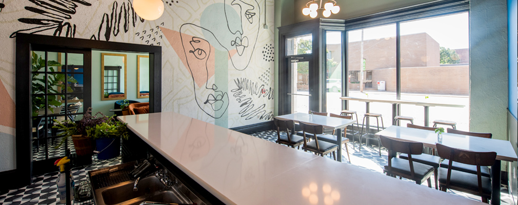 Photograph of a small restaurant with a counter in the foreground and tables and chairs in the middle of the room framed by a wall with a modern mural and another wall with the front door and large windows.