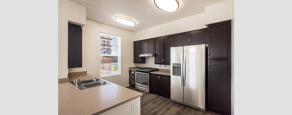 Photograph of a kitchen with a sink in a peninsula and a refrigerator, oven, and cabinets along the back wall. 