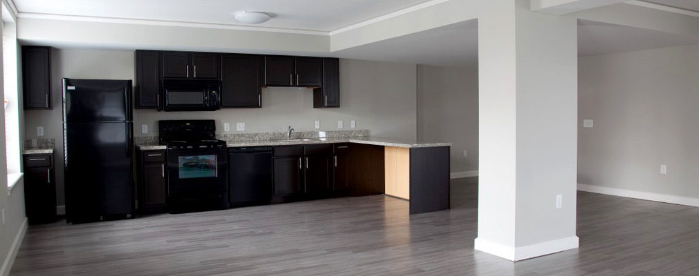 Photograph of the interior of an apartment across an unfurnished living room to a kitchen.