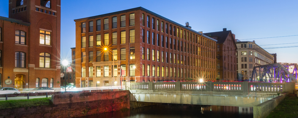 Photograph taken at twilight of two façades of a five-story brick mill building flanked by two large mill buildings beside a canal.