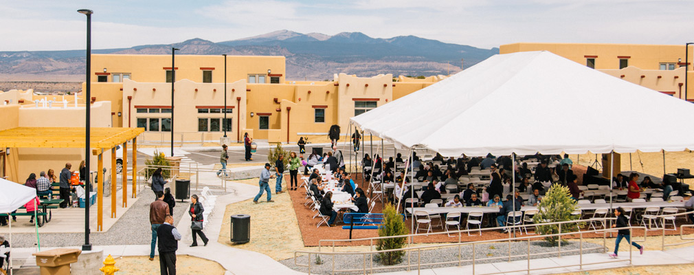 Photograph taken from one story above ground of community members standing near and seated under a large tent during the Cedar Hills’ opening ceremony. The single-story community building is at the left edge of the photograph, and a row of attached residential buildings is in the middle ground.