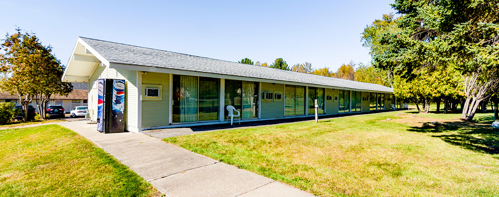 Photograph of the rear façade of a one-story, eight-unit residential building, with glass sliding doors in each unit opening to a narrow patio and lawn; the front façade of a similar building is in the background behind a parking lot.  