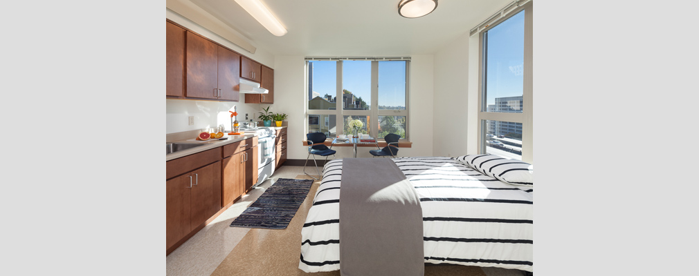 Photograph of the kitchen, sleeping, and eating areas in a studio apartment, with large windows in two walls.