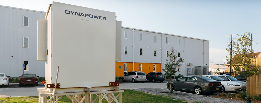 Photograph of a battery storage system in front of a parking area and a three-story apartment building.