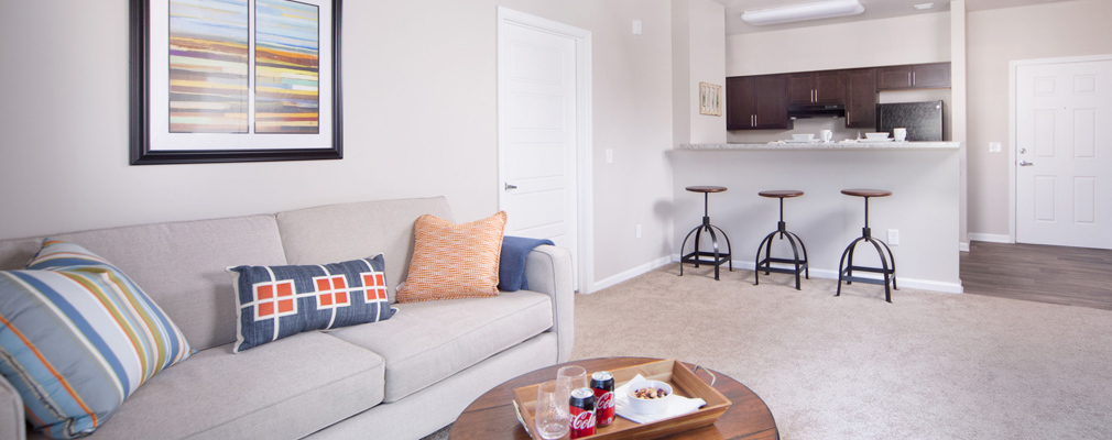 Photograph of an apartment living area with a sofa and coffee table in the foreground and kitchen area with three stools at a counter in the background. 