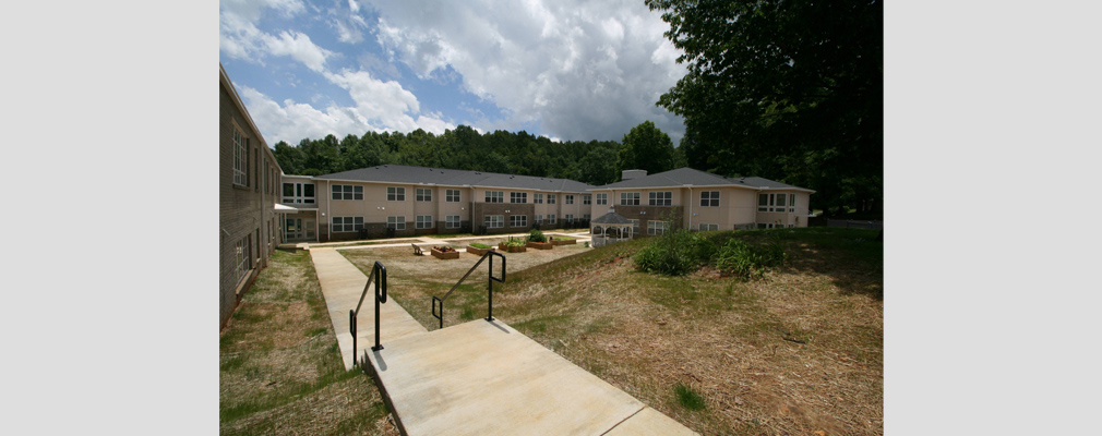 Photograph of a walkway leading to a courtyard containing five raised planters and a new, two-story building in the middle-ground.