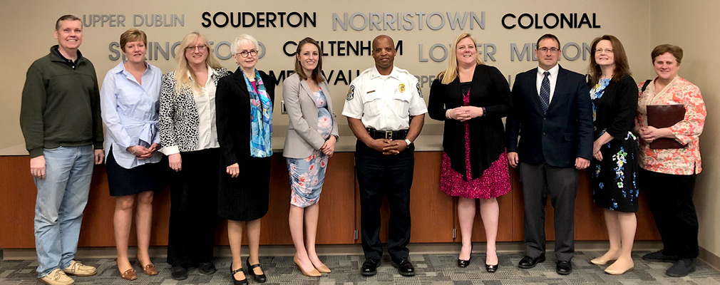 Photograph of 10 members of the Your Way Home Advisory Council posing in front of a wall adorned with the names of Montgomery County municipalities.