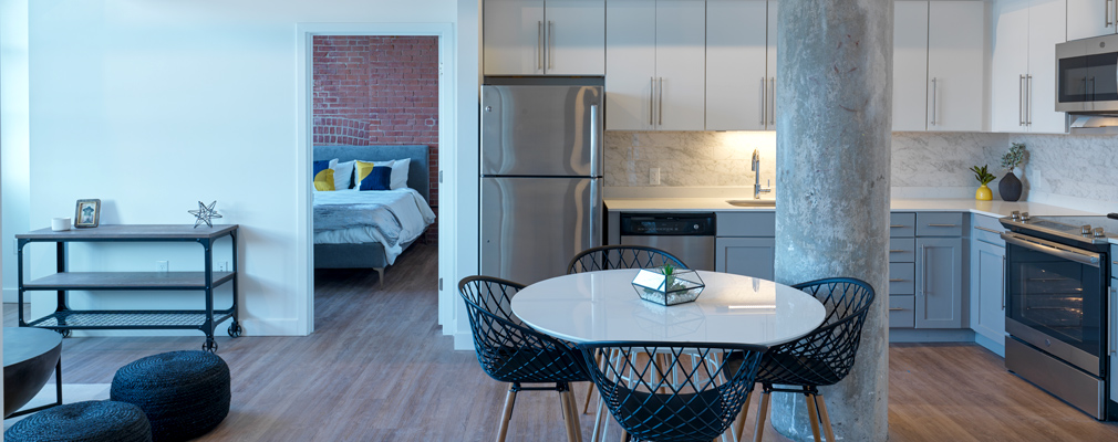 Photograph of the kitchen area of an apartment, with a sitting area to the side and a doorway leading to a bedroom.