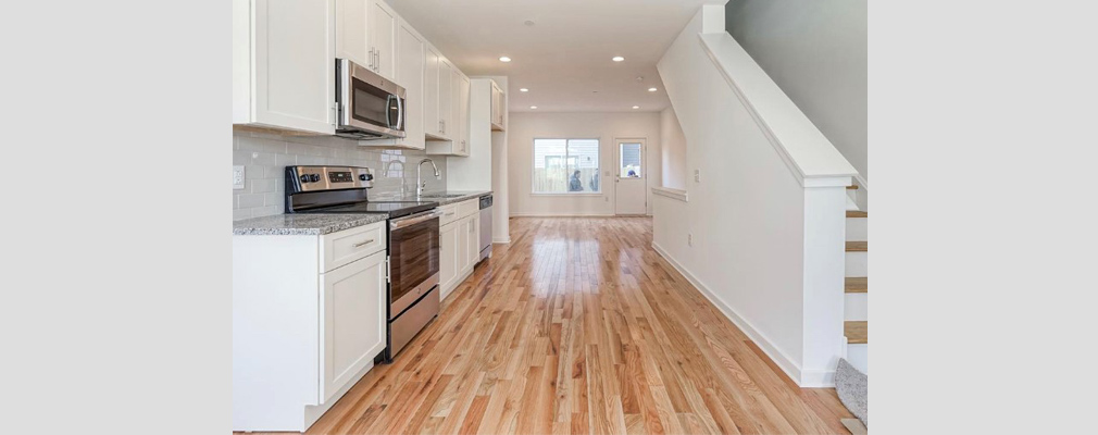 Photograph of a kitchen with hardwood floors and solid wood cabinets and a staircase leading to an upper floorin the foreground and, in the background, a living area with the front door and a large window in the far wall.