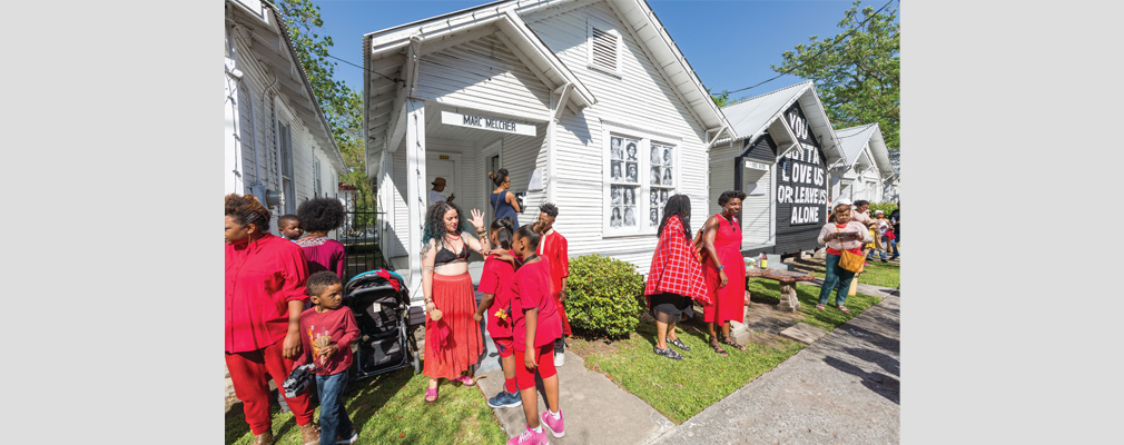 Photograph of women and children gathering on the front lawns of several shotgun houses with artwork on the front facades.