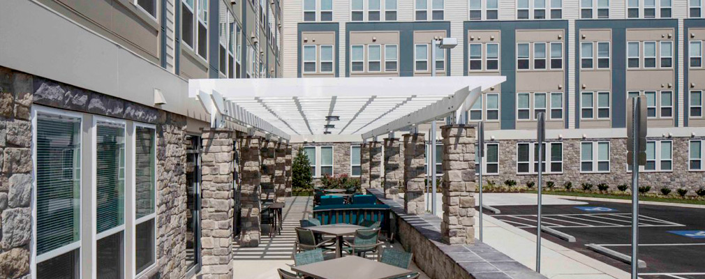 Photograph of a patio with a pergola shading several tables and chairs beside a multistory apartment building.