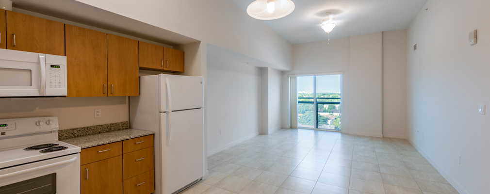 Photograph of the interior of an unfurnished apartment, with a kitchen area in the foreground, a large living area in the middleground, and a sliding glass door in the far wall.