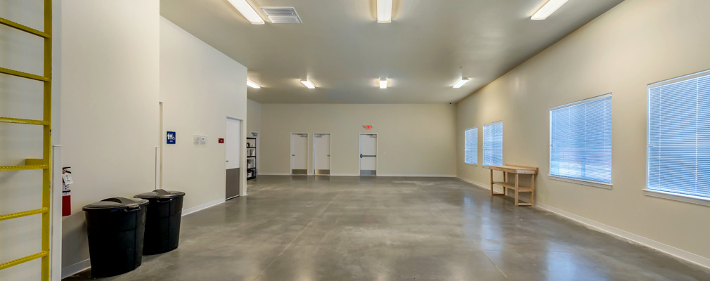 Photograph of a large, unfurnished space with a concrete floor, fluorescent lights, and large windows. 