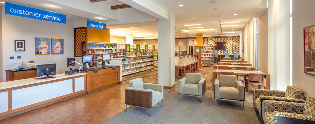 Photograph of the information desk and a sitting area in the foreground, with book stacks, tables, and computer stations in the background.