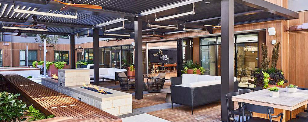 Photograph of a trellis-covered courtyard with tables, seating, an outdoor fireplace, and landscaping. 