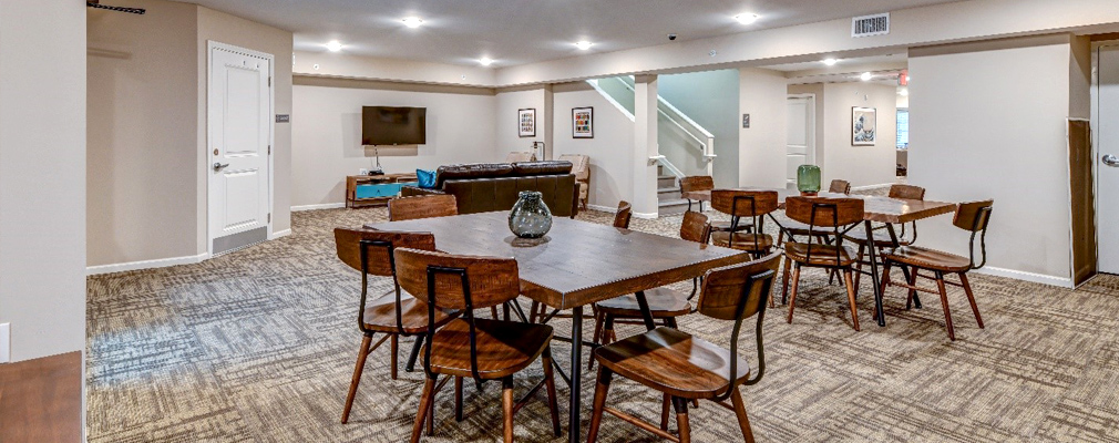 Photograph of a large room with two tables and several chairs in the foreground and a sofa and television at the back wall. 