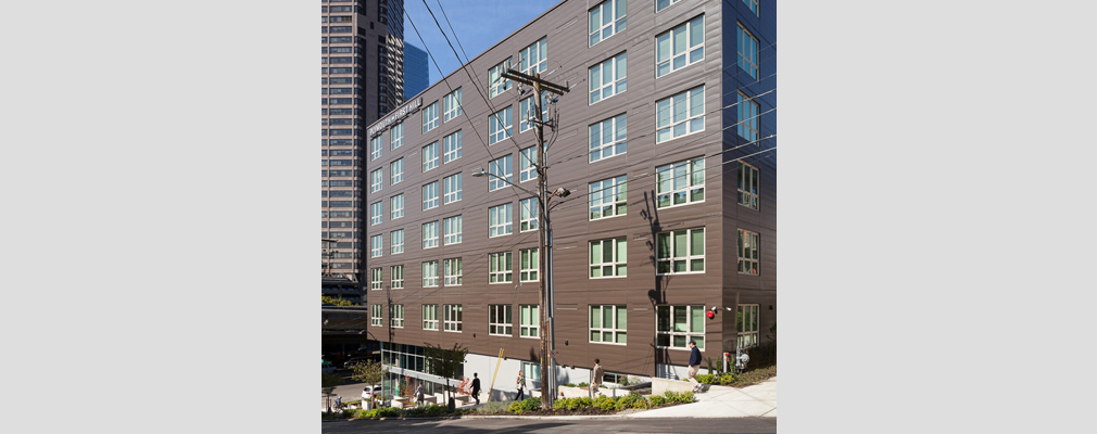 Photograph of the side façade of a six-story apartment building, with two office towers in the background.