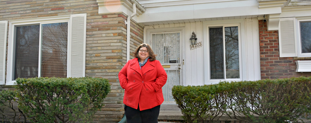 Photograph of a woman standing in front of a one-story brick single-family house. 