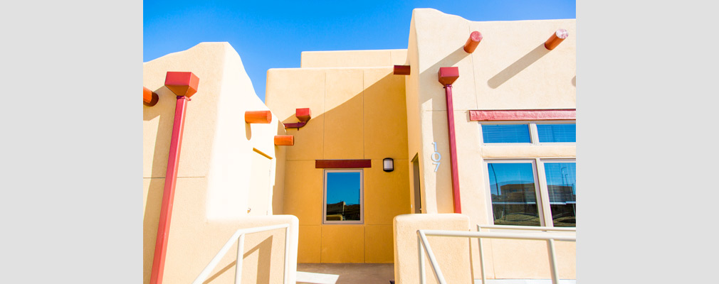 Photograph of the front façade of an attached dwelling, with a stucco veneer and vigas as well as glazed windows, handrails, and downspouts.