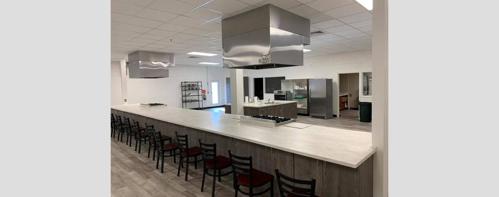 Photograph of a large commercial kitchen space with an observation counter set with two ranges and chairs in the foreground and additional kitchen appliances in the background.