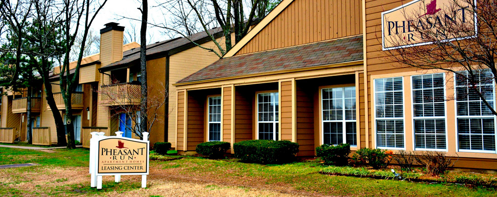 Photograph of a two-story apartment building with an adjacent leasing center and a sign in the foreground reading “Pheasant Run Apartment Homes Leasing Center.”