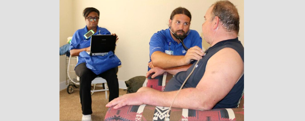 Photograph of two health workers delivering a checkup to a patient in his living room.