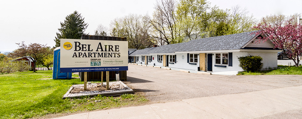 Photograph of the front façade of a single-story, eight-unit residential building under construction, with a sign in the foreground reading “Coming Soon: Bel Aire Apartments, a collaboration of Champlain Housing Trust and the University of Vermont Medical Center.” 