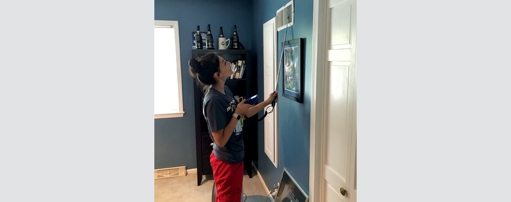 A woman measures airflow at a living room air vent.