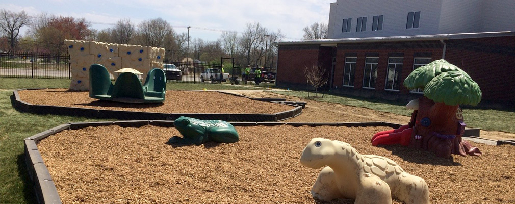 Photograph of a mulched playground featuring plastic animals in the foreground, with the rear of the two-story educational building in the background on the right side of the photograph.