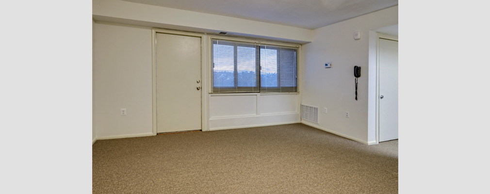 Photograph of a corner of an unfurnished living room, with the front door and a set of windows along one of the walls.