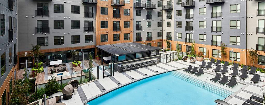 Photograph of a courtyard surrounded by a five-story building and featuring a swimming pool, a sitting area covered with a pergola, and an uncovered sitting area with an outdoor fireplace.