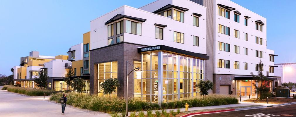 Photograph from the street of a multistory residential building, with a community room in the foreground and an entrance to the building at the right edge of the photograph. 