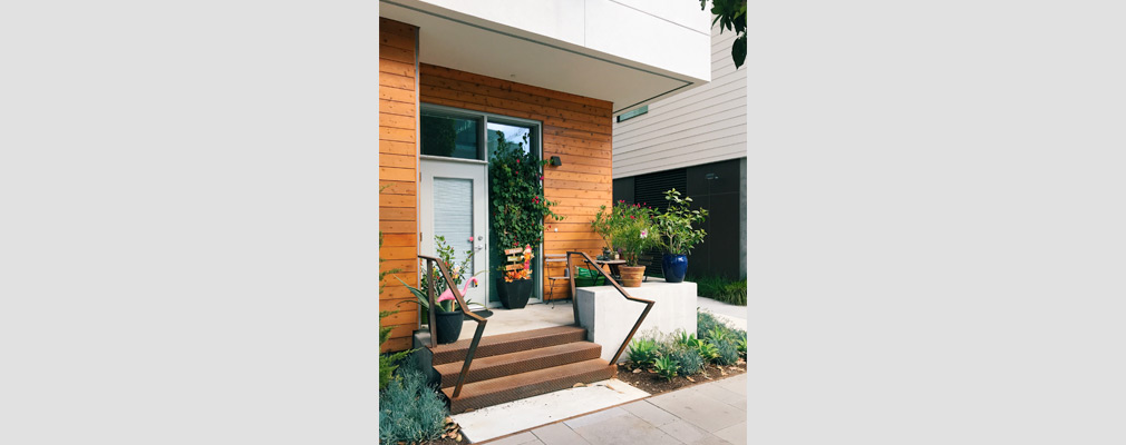 Photograph of a stoop at the front door of an apartment, raised three steps above the sidewalk and decorated with a table and chair, plants, and a plastic flamingo.