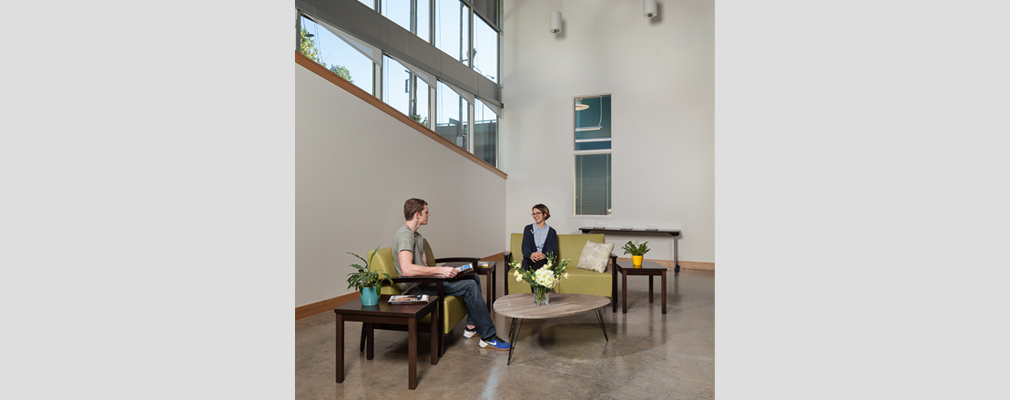 Photograph of two people sitting on upholstered loveseats near several tables in a large room with clerestory windows in a side wall.