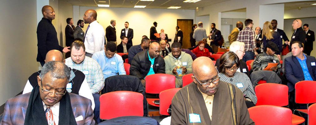 Photograph of several dozen people seated and standing in a large meeting room. 