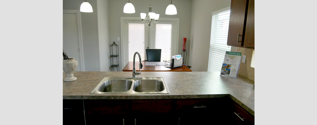 Photograph taken from behind a kitchen peninsula with a sink into a dining area with a large window and two glass doors.