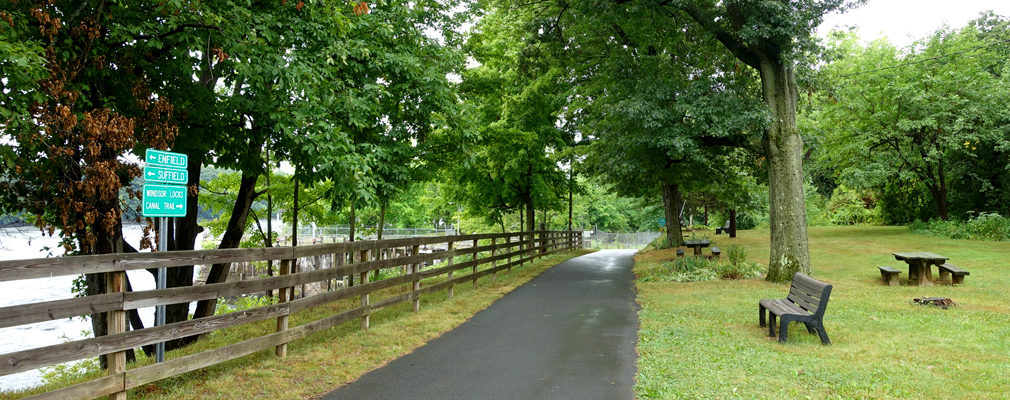 Photograph of a paved trail running beside a river, with a rail fence, bench, and picnic table near the trail.