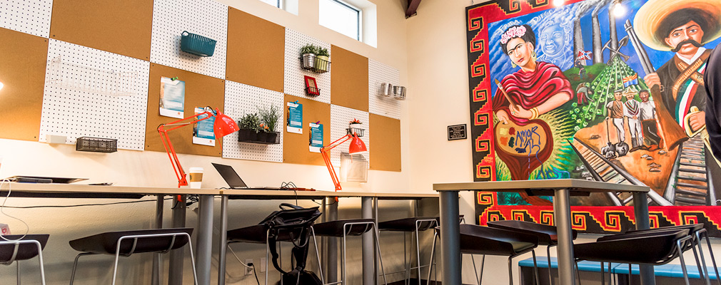 Photograph of a corner of a classroom with a Latino-themed mural on one wall and desks along another wall with pegboard and cork tiles and clerestory windows.