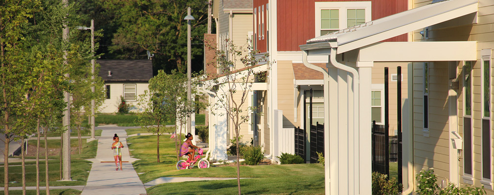 Photograph of a tree-lined sidewalk where two young girls play in front of a row of houses, several of which have porches.
