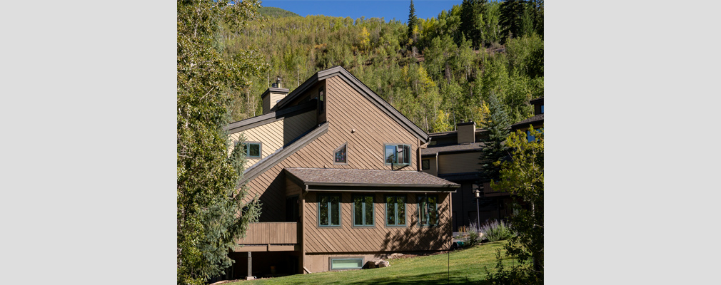 Photograph of a side facade of a three-story townhouse against a forested mountain backdrop. 