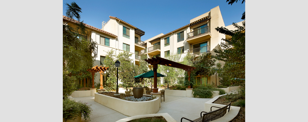 Photograph of a paved area with pergolas and seating within a landscaped courtyard framed by a multistory building.