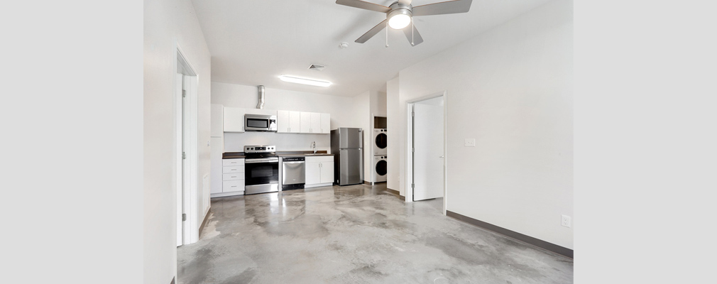 Photograph taken from the living area into a kitchen area with upper and lower cabinets and a stove, microwave oven, dishwasher, refrigerator, and clothes washer/dryer.