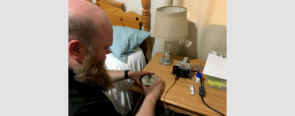Photograph of a man in front of a bedside nightstand handling air-testing equipment.