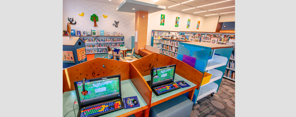 Photograph of two computers for children, with a sitting area and book stacks in the background.